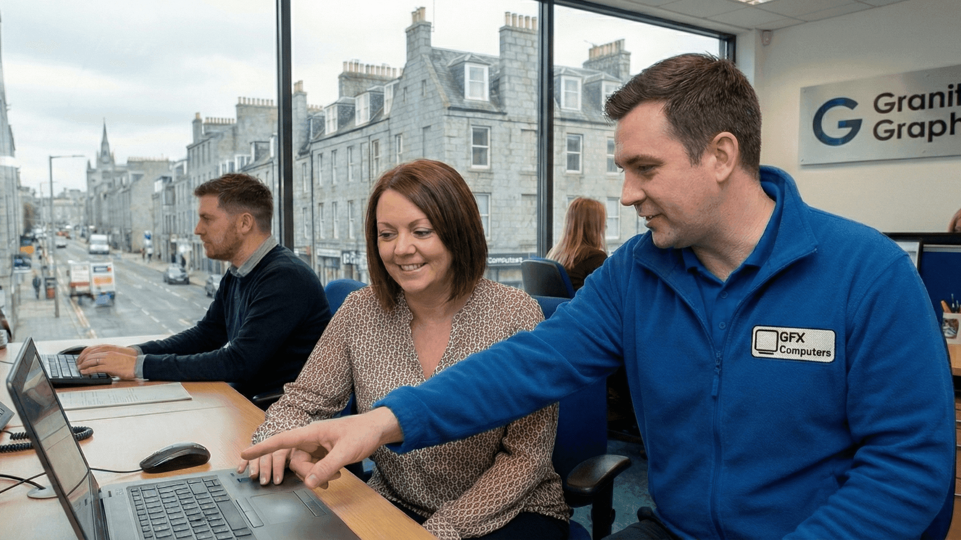 GFX Computers engineer providing friendly IT support to a client in an Aberdeen office, with city buildings visible through the window.